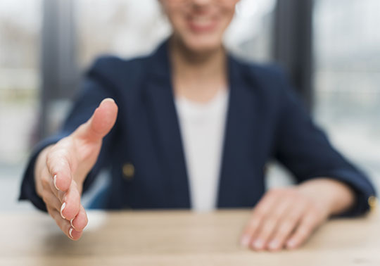 Bewerberin im blauen Anzug sitzt an einem Tisch und streckt lächelnd die rechte Hand zur Begrüßung aus